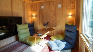 A cozy room with wooden walls, a flat-screen TV, a small table with a vase, and two cushioned seating areas with pillows. - JR Mobile Inn Sapporo Kotoni (Sapporo)