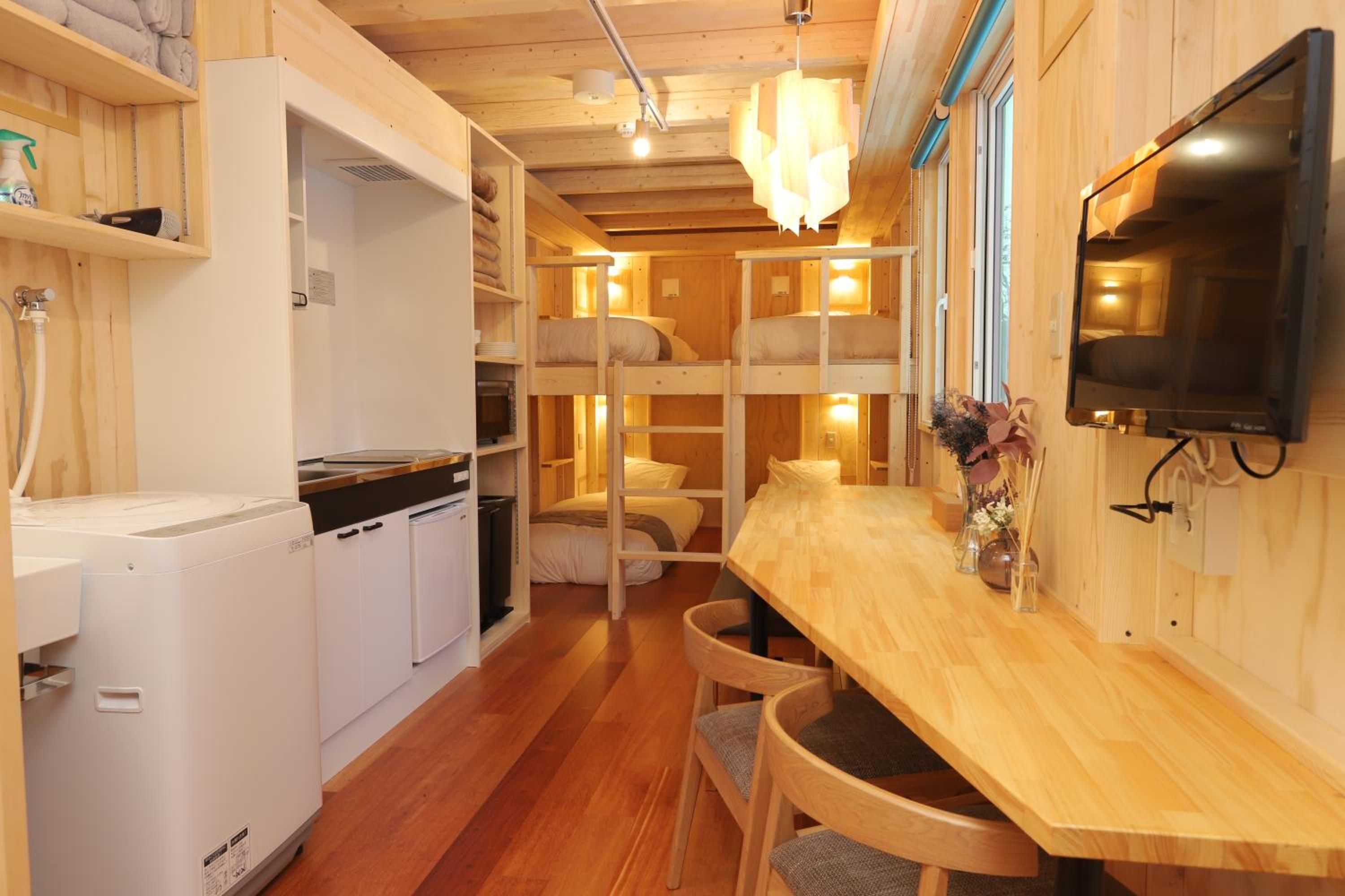 A compact kitchen with a washing machine, a dining area with a wooden table and chairs, and a television mounted on the wall.