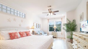 A bedroom with a large bed, a ceiling fan, a window with curtains, a nightstand, a dresser, and a plant. - 3BR/3BA Oceanview just steps from the sand~ 107 Ocean Pointe (Folly Beach)