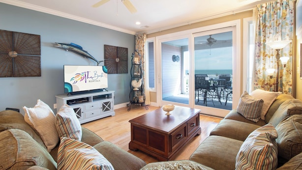 A living room with a large sofa, a coffee table, and a television. There is a ceiling fan, a window with curtains, and a view of the outside. - 3BR/3BA Oceanview just steps from the sand~ 107 Ocean Pointe (Folly Beach)