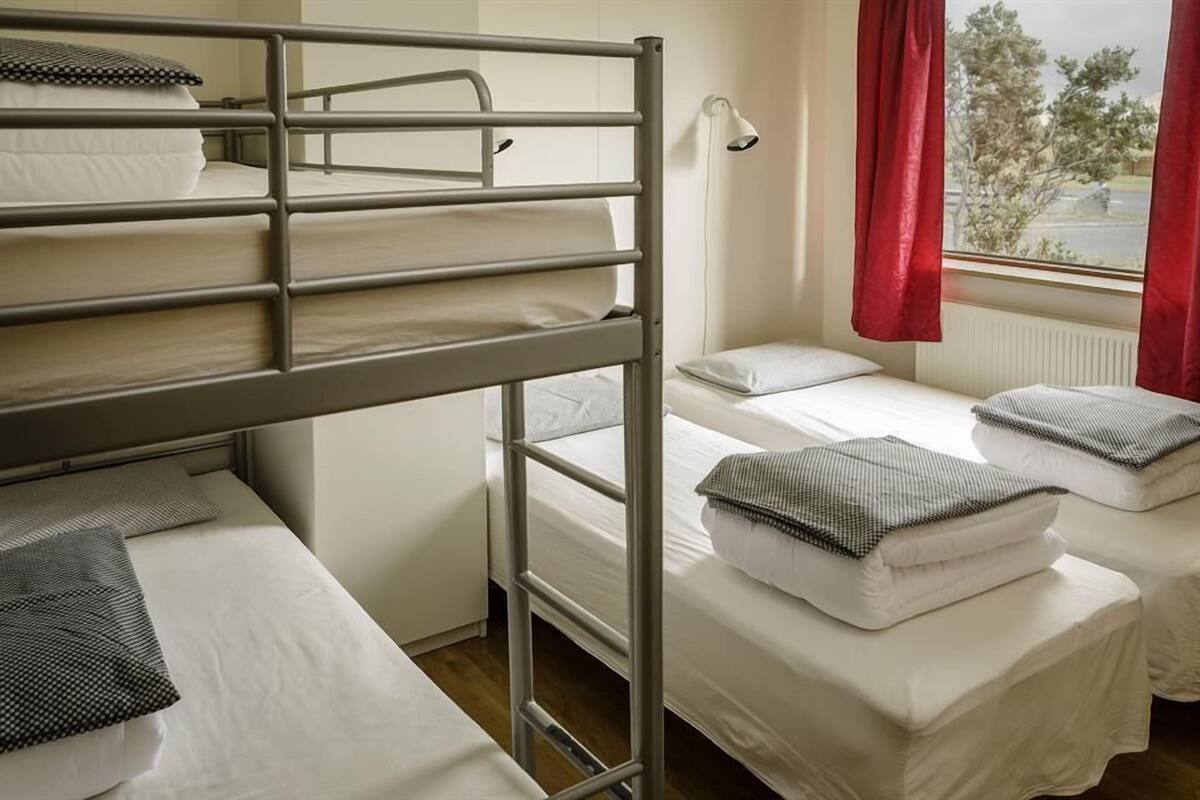A dormitory room with bunk beds, a desk, and a window with red curtains.