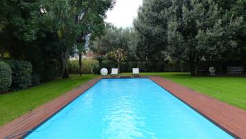 Ein rechteckiger Pool mit einer Holzterrasse und einer Rasenfläche mit Bäumen und Stühlen.
