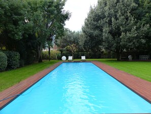 Une piscine rectangulaire dotée d’une terrasse en bois et d’une zone herbeuse agrémentée d’arbres et de chaises.