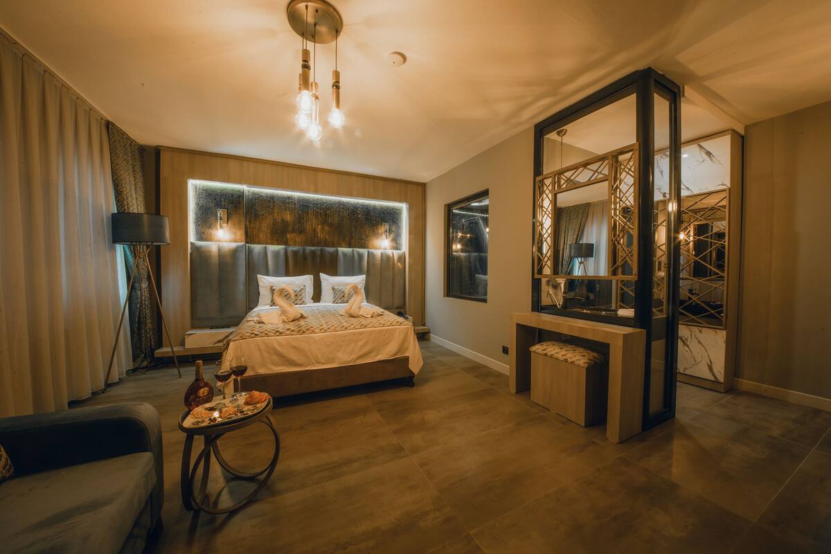 A modern hotel room with a large bed, a bedside table with a bottle and glasses, a small round table with a tray, and a large mirror.
