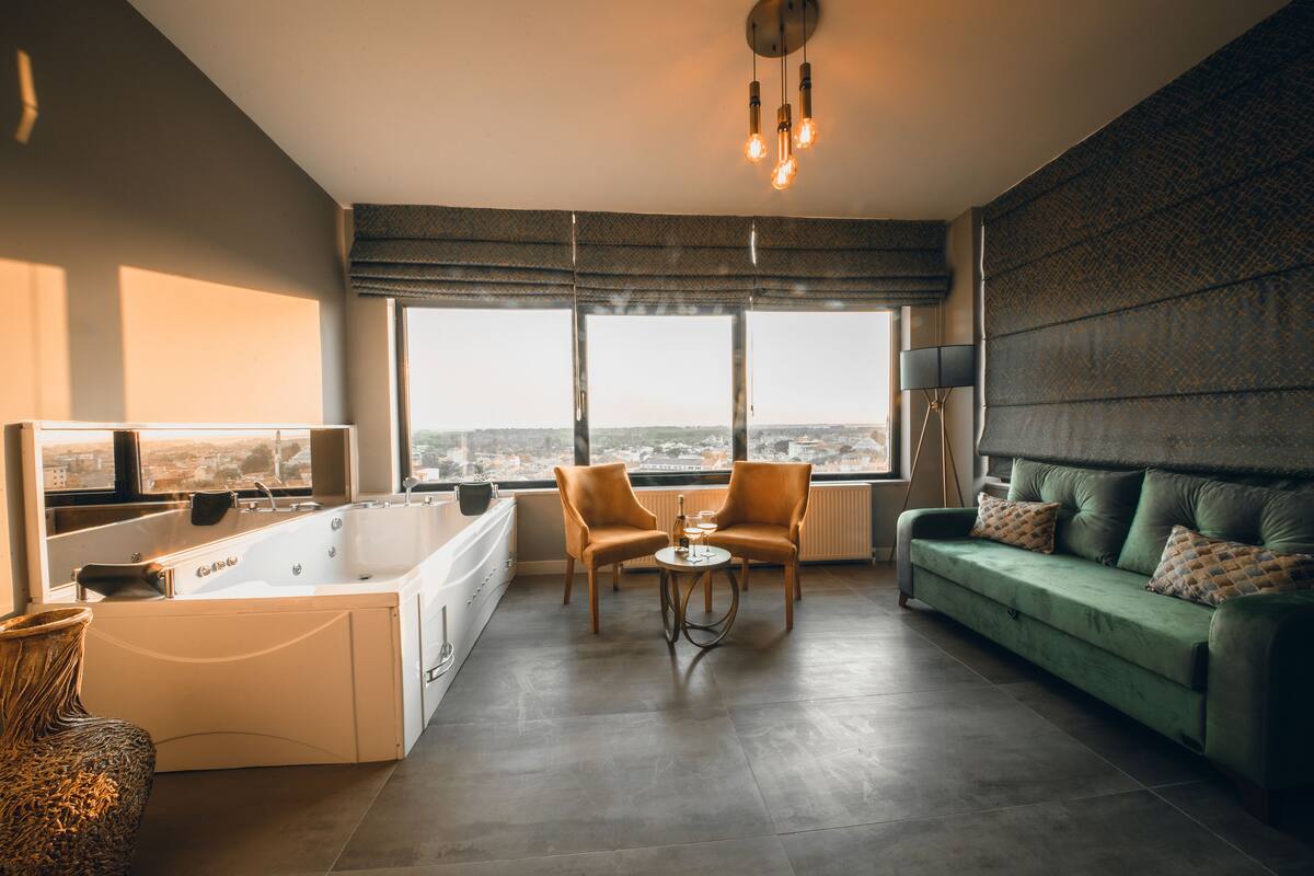 A modern living room with a large window, a green sofa, two armchairs, a small round table, and a bathtub.