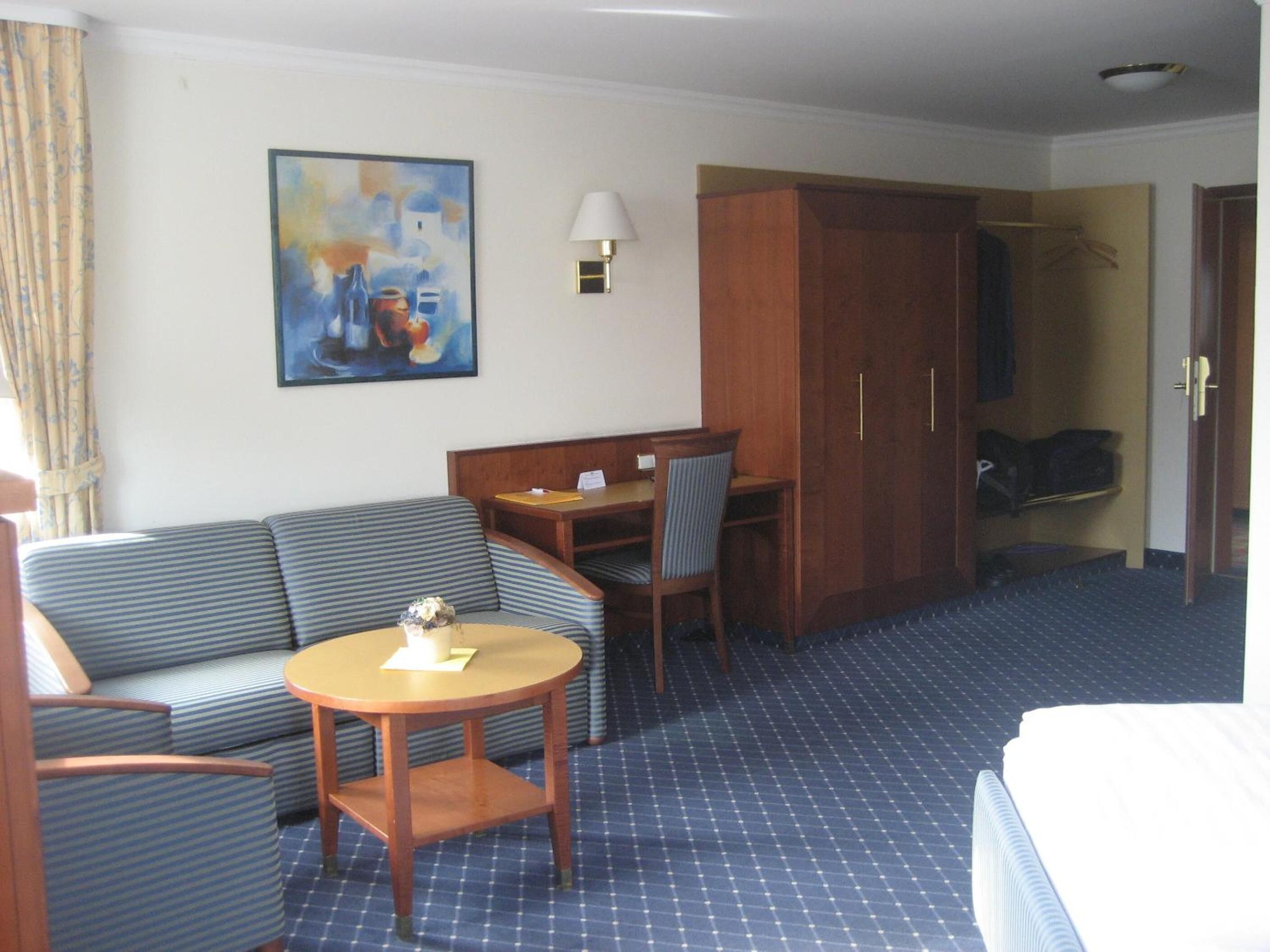 Ein Hotelzimmer mit einem Bett, einem Sofa, einem kleinen Tisch, einem Sessel, einem Schreibtisch, einem Kleiderschrank und einem Bild an der Wand.