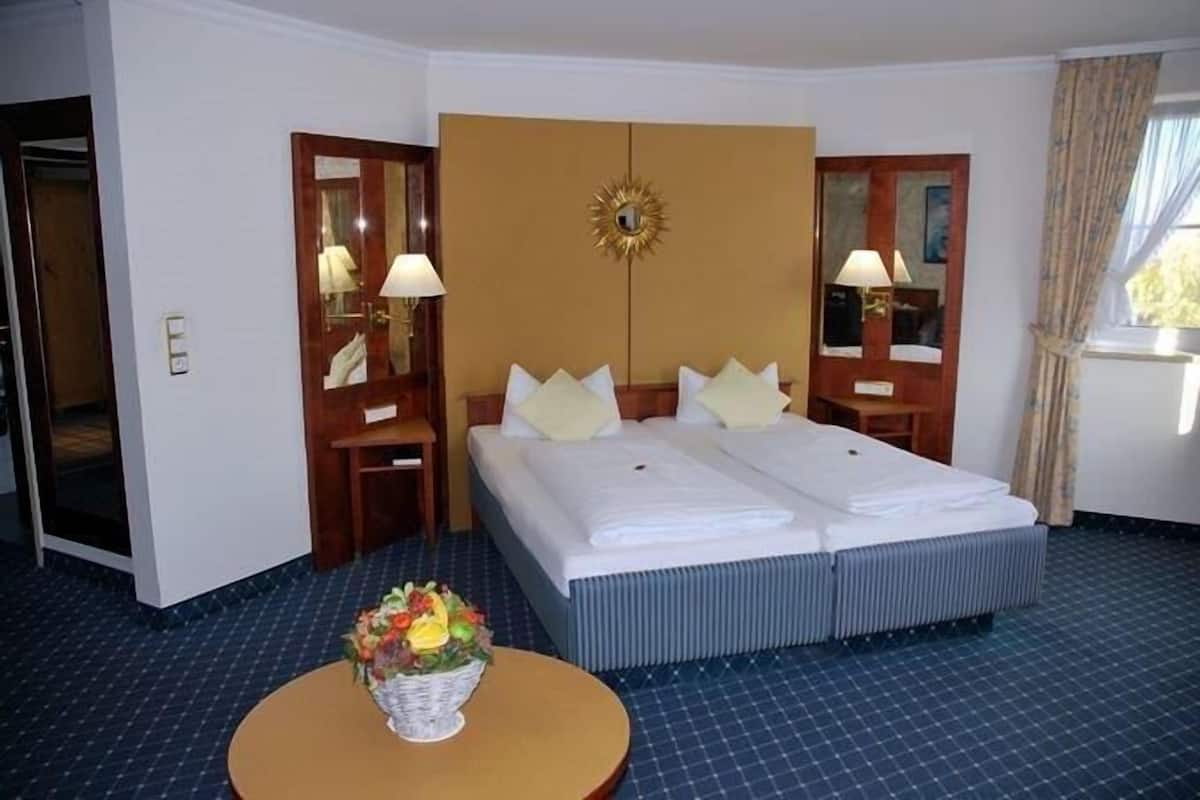 Habitación de hotel con cama doble, dos mesitas de noche con lámparas, un espejo grande y una pequeña mesa redonda con un arreglo floral.