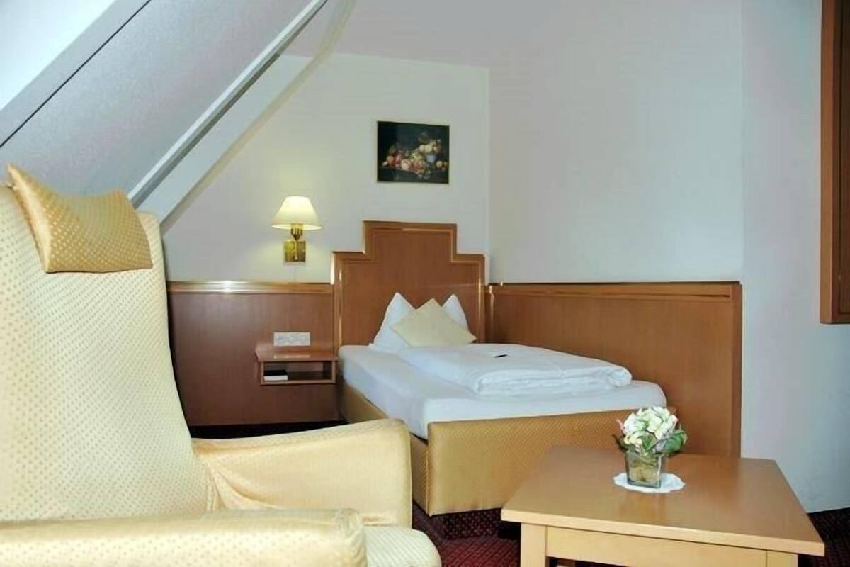 Habitación de hotel con una cama, una silla, una mesita con un florero y una mesita de noche con una lámpara, además de un cuadro en la pared.