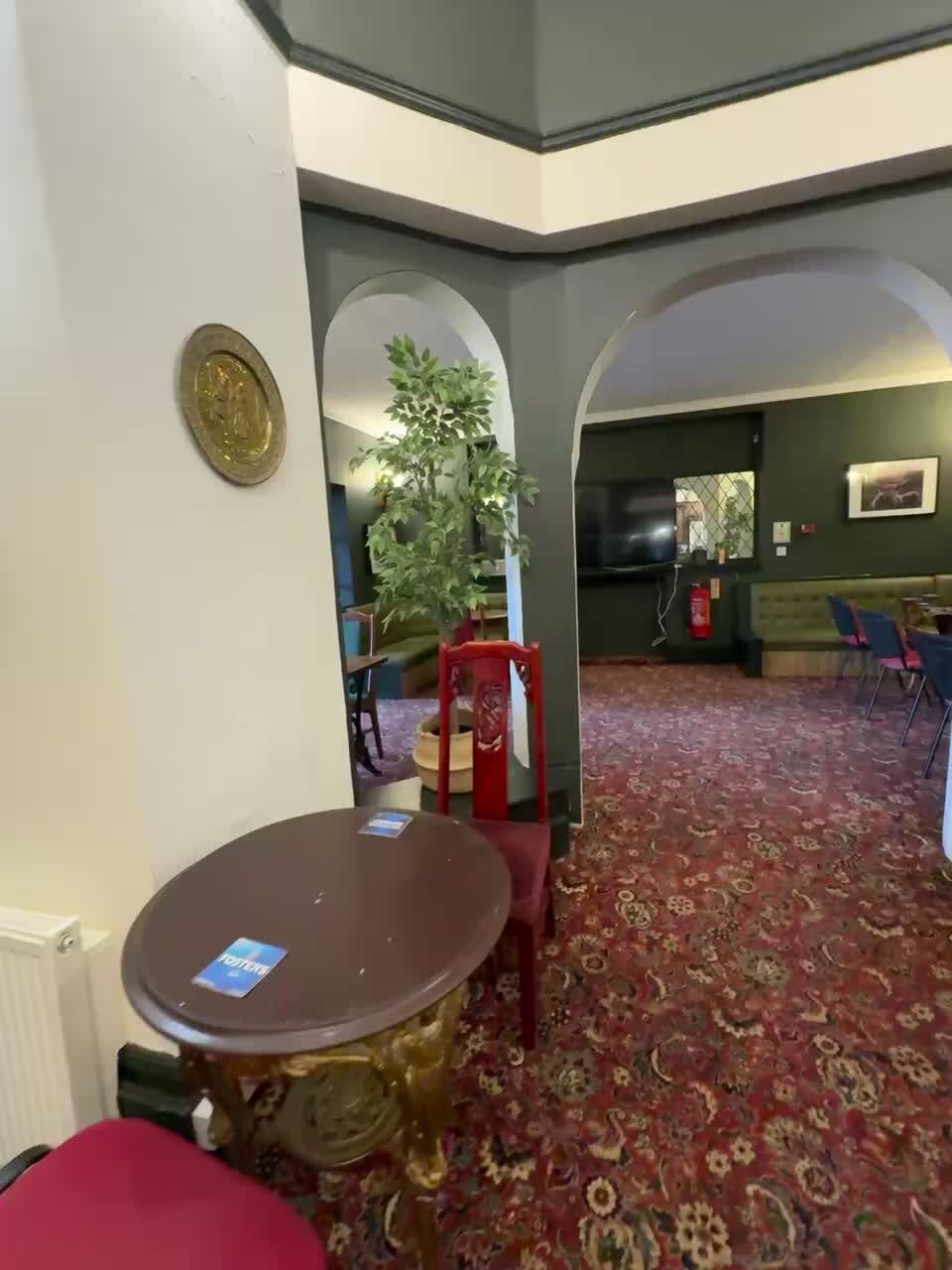 A hotel lobby with a round table, red chairs, a potted plant, and a patterned carpet.