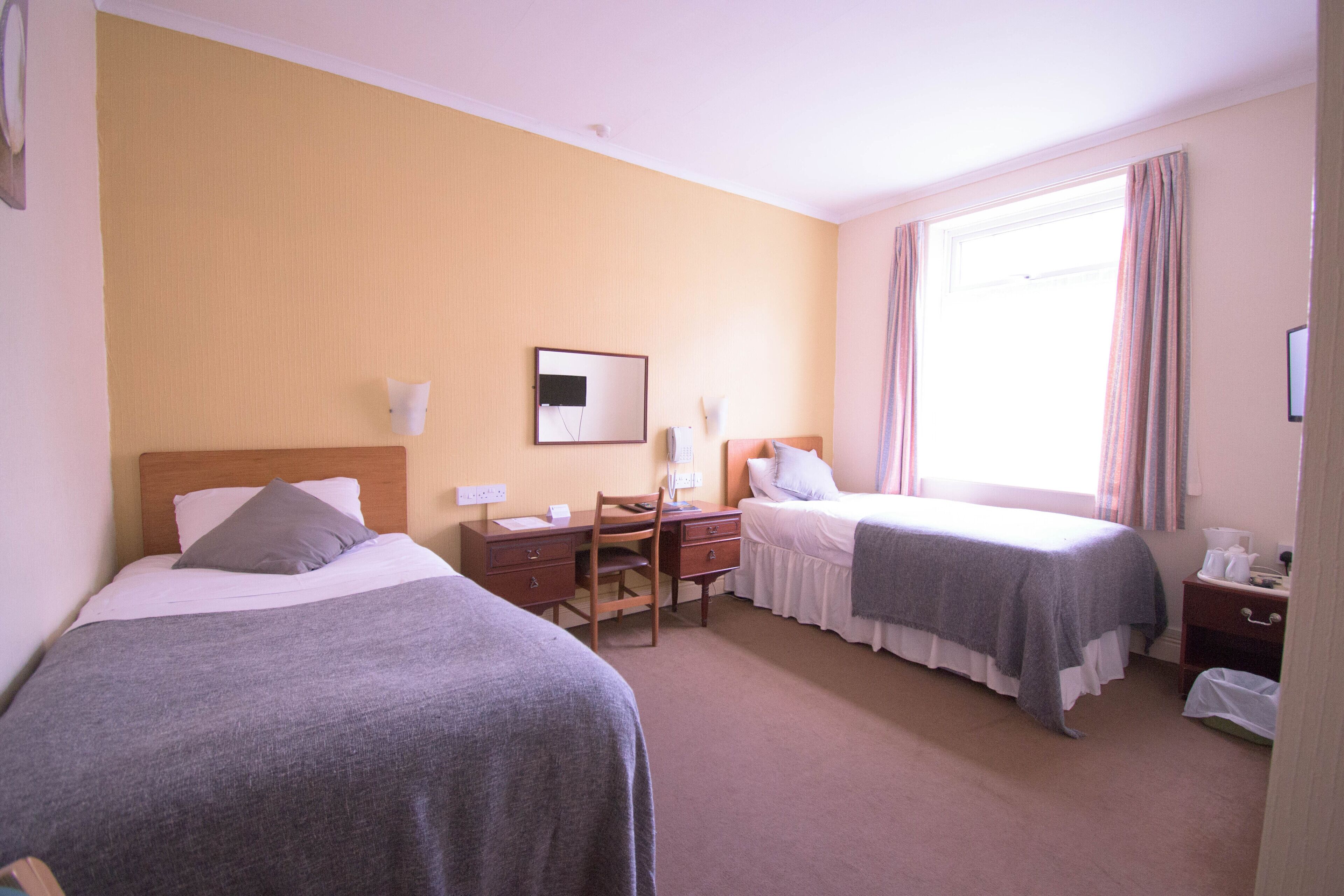 Une chambre d’hôtel avec deux lits simples, un bureau avec une chaise, une petite table et une fenêtre avec des rideaux.