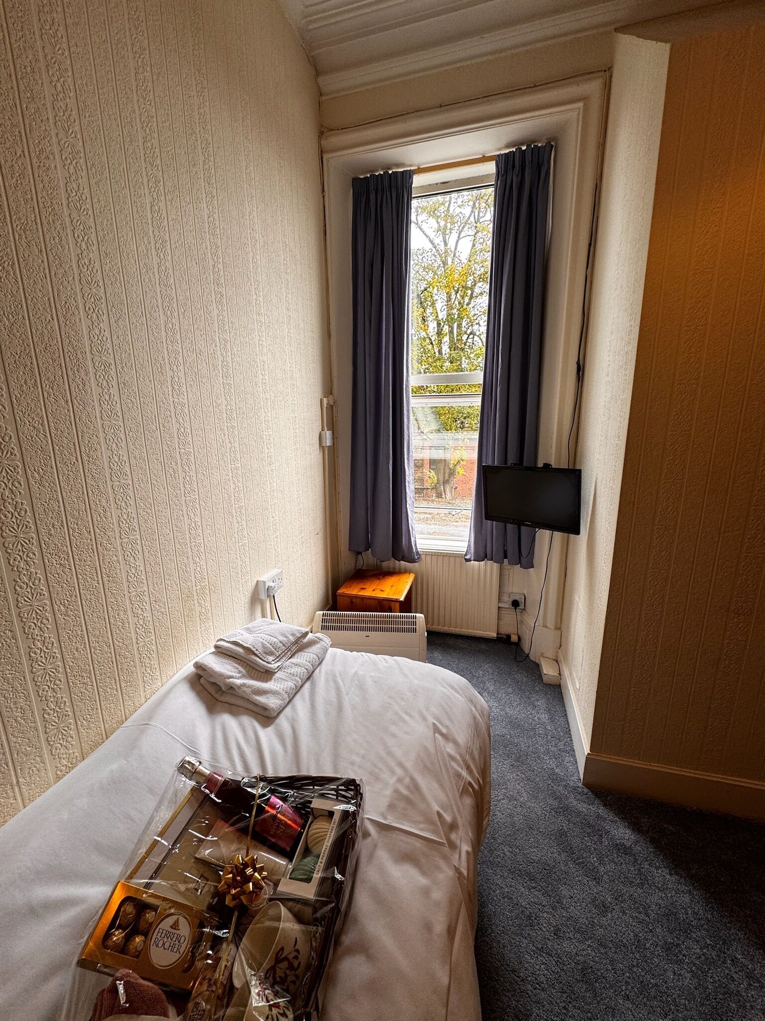 Quarto de hotel com uma cama, uma mesinha com artigos de higiene, uma TV fixada na parede e uma janela com cortinas.