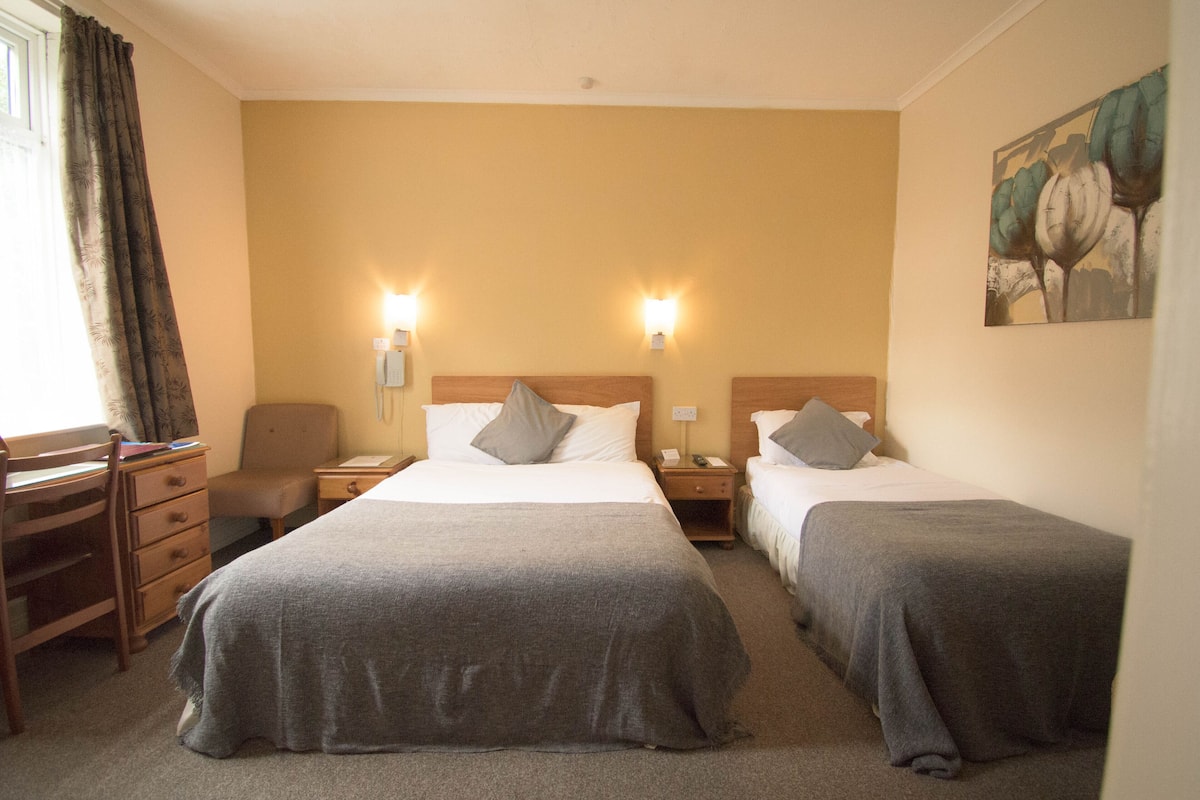 Quarto de hotel com duas camas de solteiro, uma escrivaninha, uma cadeira e uma mesa de cabeceira com abajur.