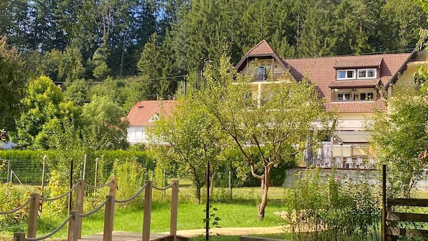 Een houten bruggetje naar een huis met rode dakpannen, omringd door weelderige begroeiing en bomen.