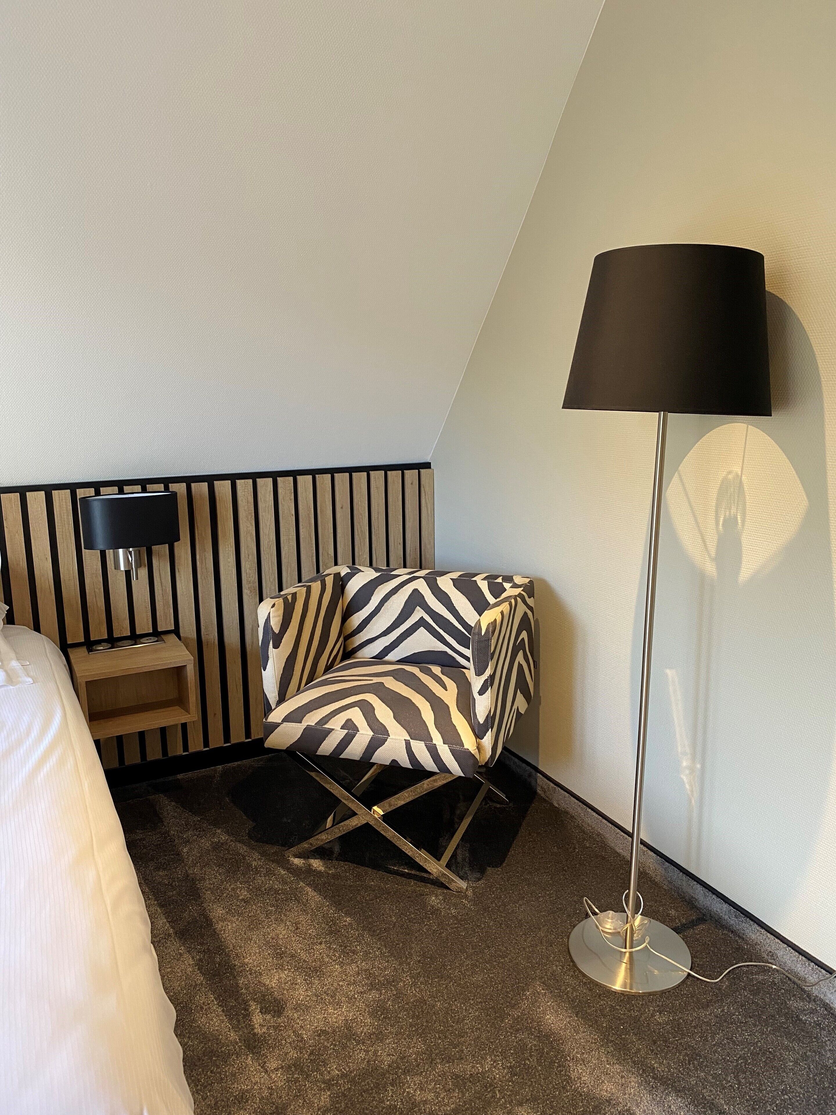 A modern hotel room with a zebra-patterned armchair, a standing floor lamp, a bedside table with a lamp, and a bed with white bedding.
