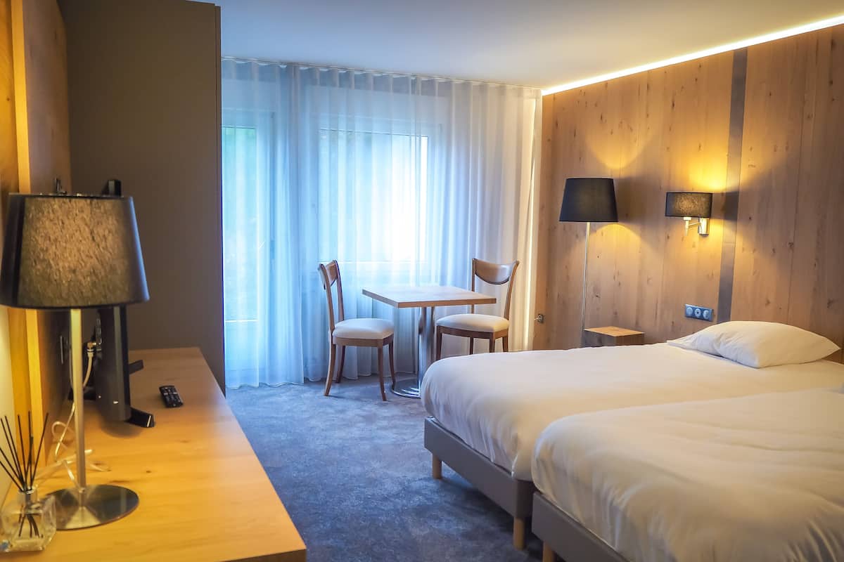 A hotel room with a bed, a desk with a lamp, a chair, a small table, and a window with curtains.