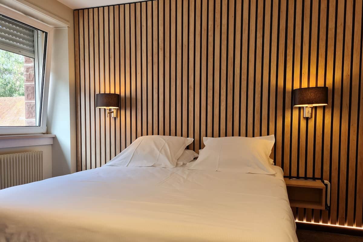 A bed with white linens, a window with blinds, and a wall with vertical wooden slats.