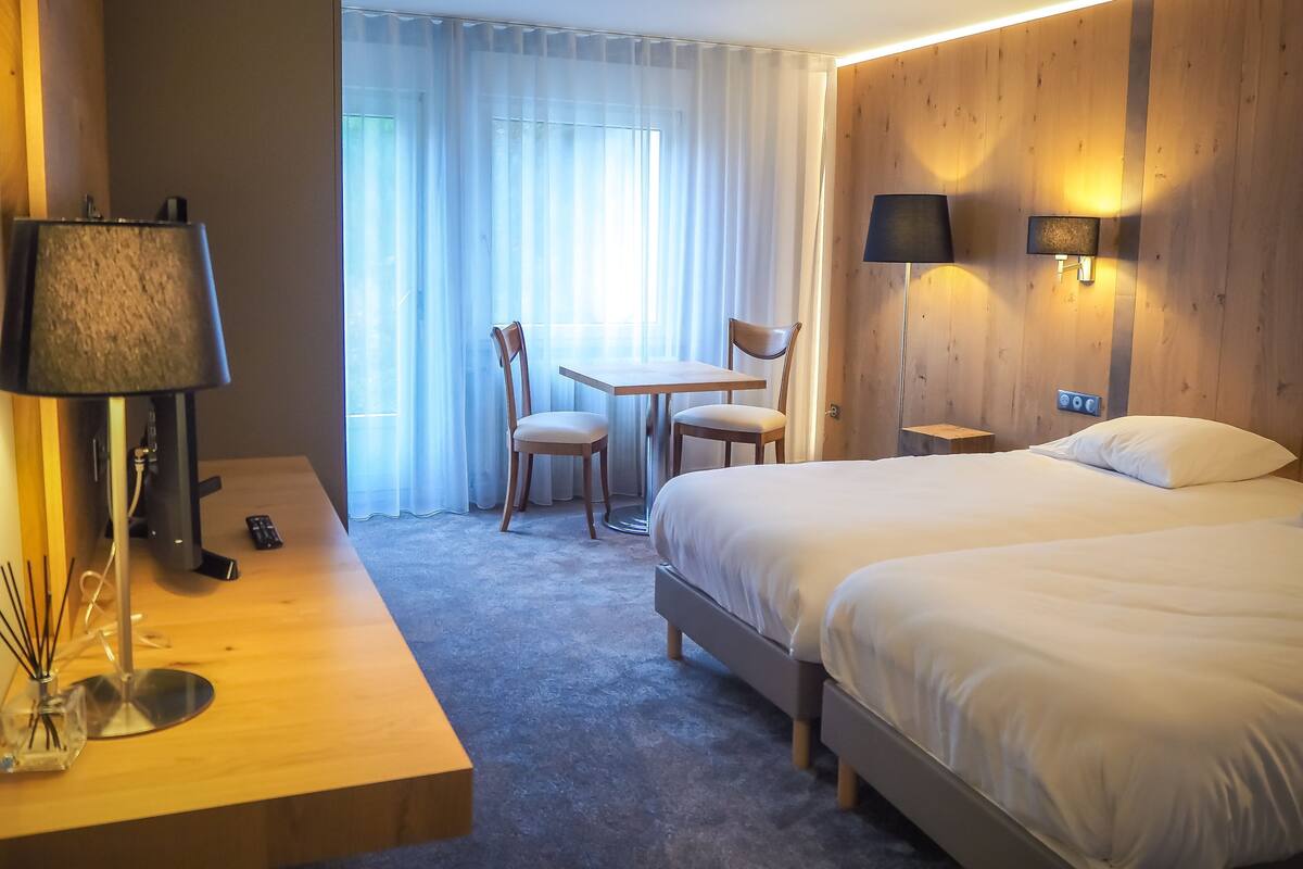 A hotel room with a bed, a desk with a lamp, a chair, a table, and a window with curtains.