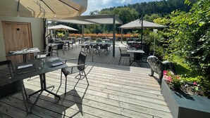 Een terras buiten met tafels en stoelen, overdekt met parasols, omringd door groen en bloemen.