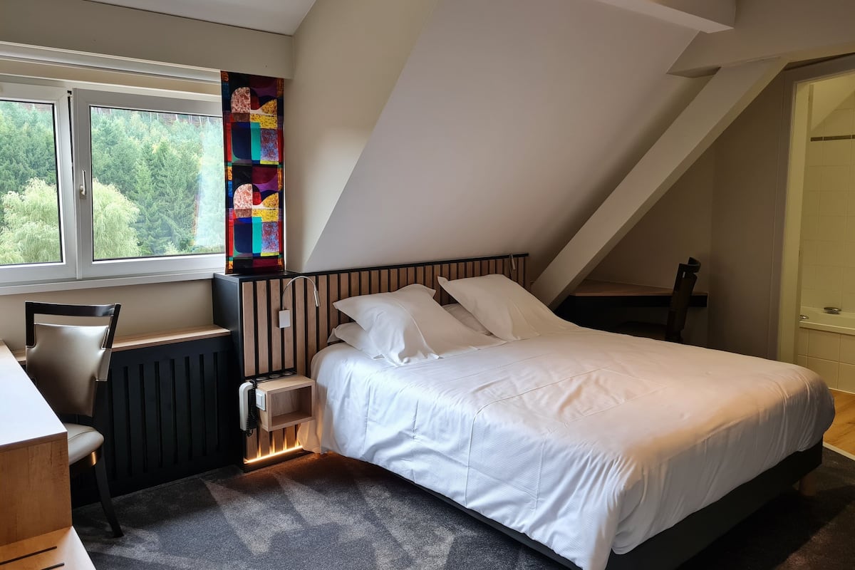 A bedroom with a sloped ceiling, a bed with white bedding, a nightstand, a chair, a radiator, and a colorful abstract artwork on the wall.