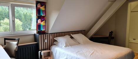 A bedroom with a sloped ceiling, a bed with white bedding, a nightstand, a chair, a radiator, and a colorful abstract artwork on the wall.