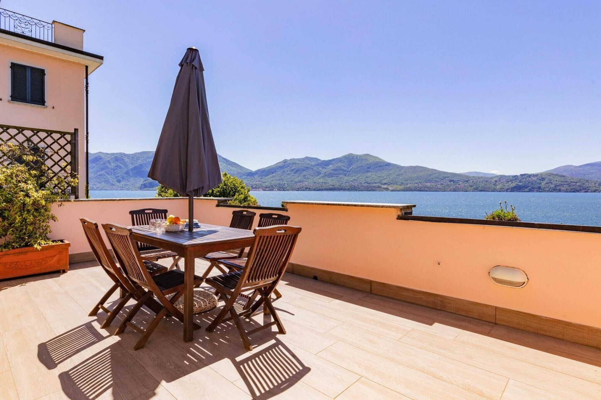 Una terraza con una mesa y sillas, un paraguas grande y vistas a un lago con montañas al fondo.