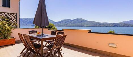 A terrace with a table and chairs, a large umbrella, and a view of a lake with mountains in the background.