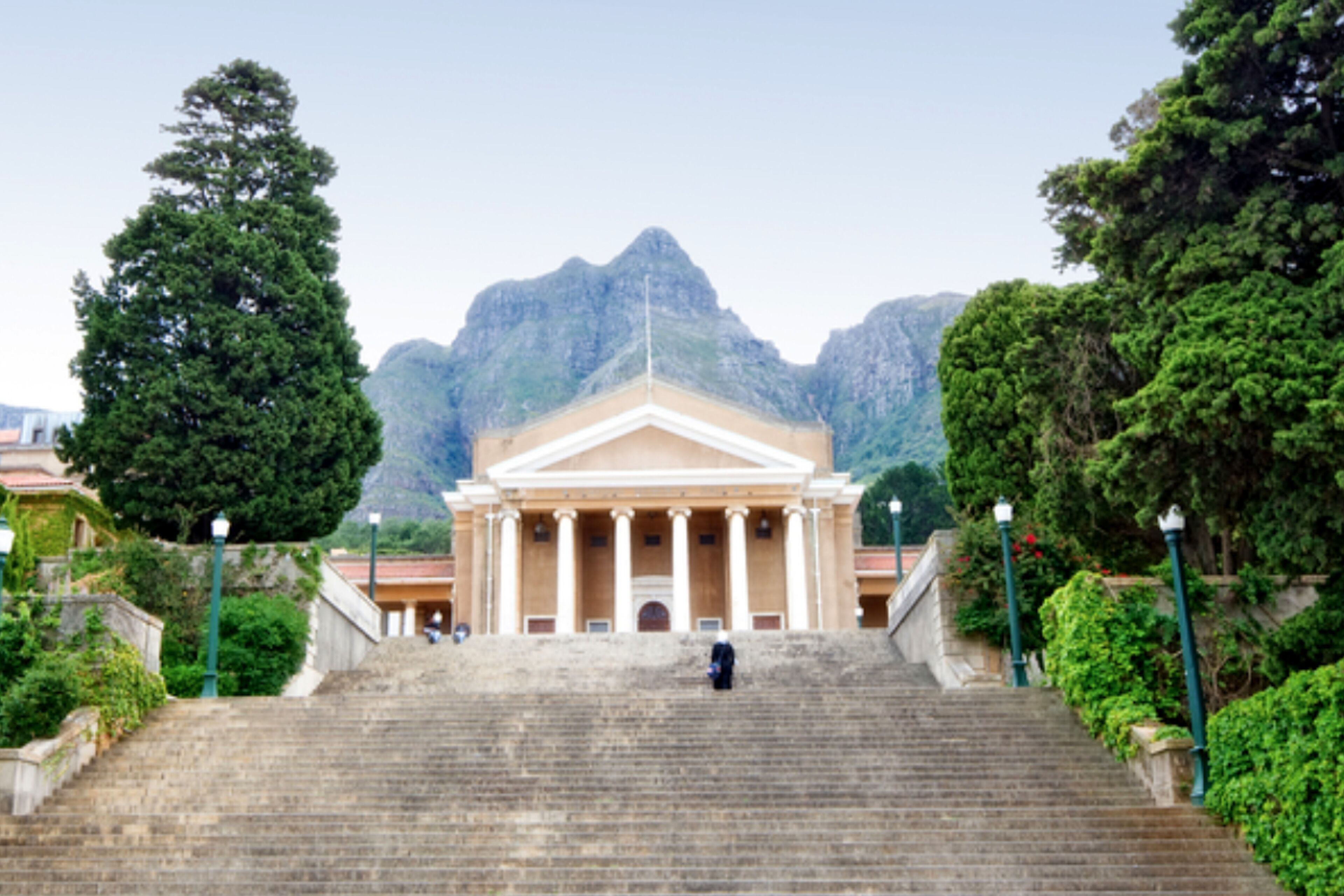 A grand building with a symmetrical facade and columns, surrounded by lush greenery and a mountain backdrop.