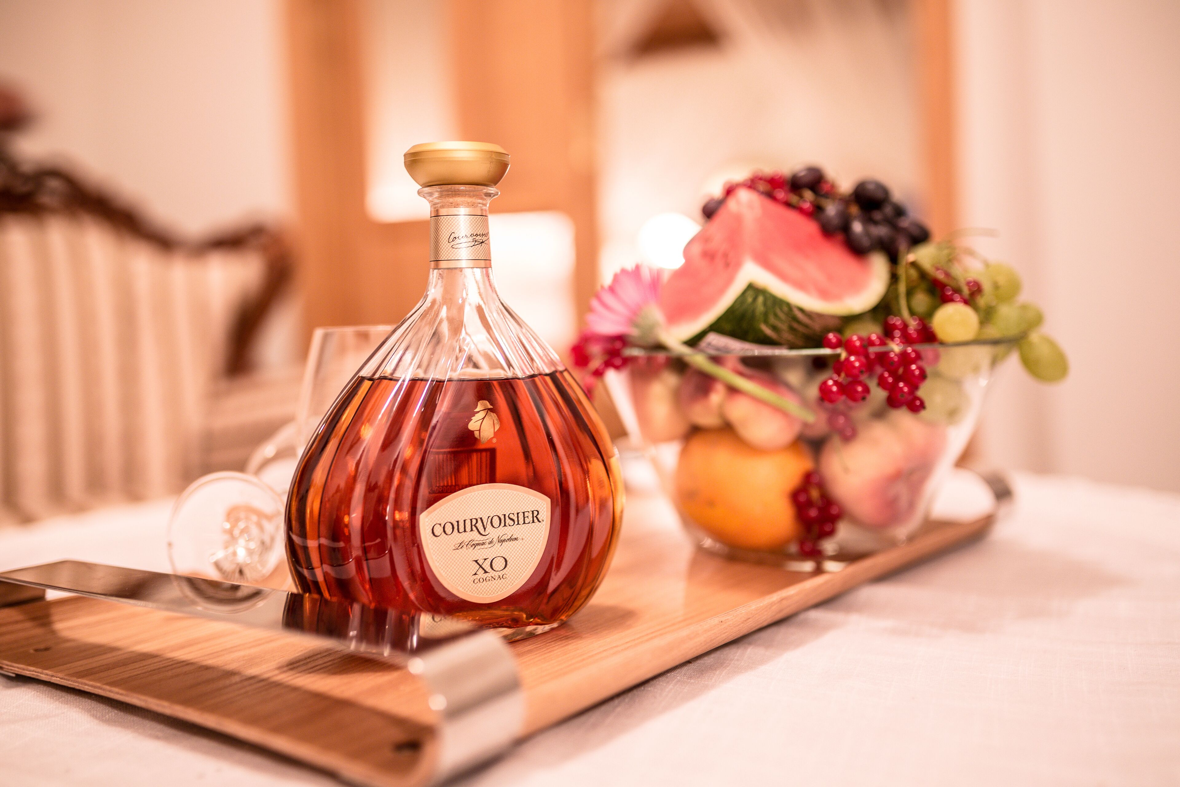 Una botella de Courvoisier XO sobre una bandeja de madera, junto a un vaso y un cuenco con fruta.