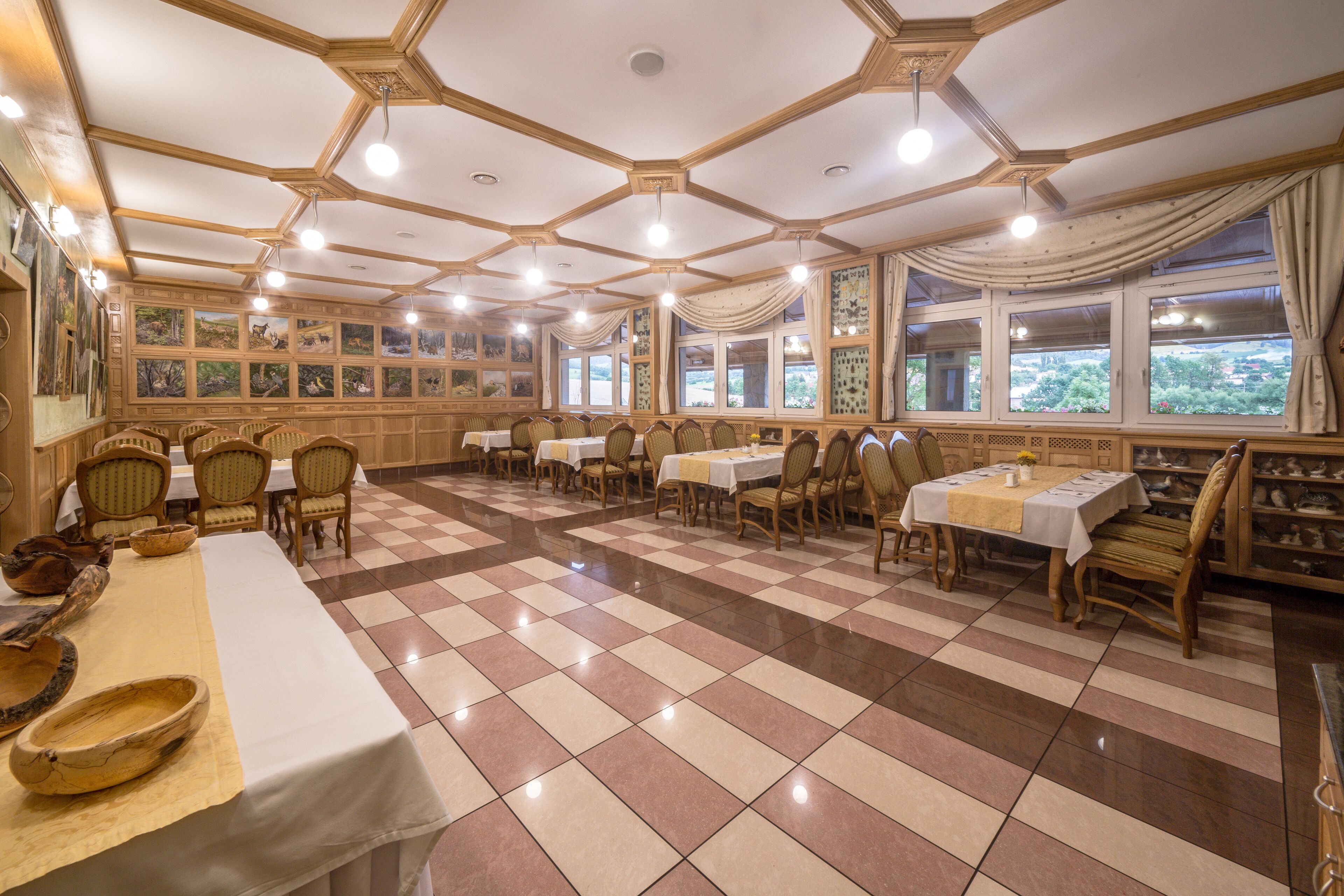 A dining area with checkered flooring, wooden chairs, tables with white tablecloths, and large windows with curtains.