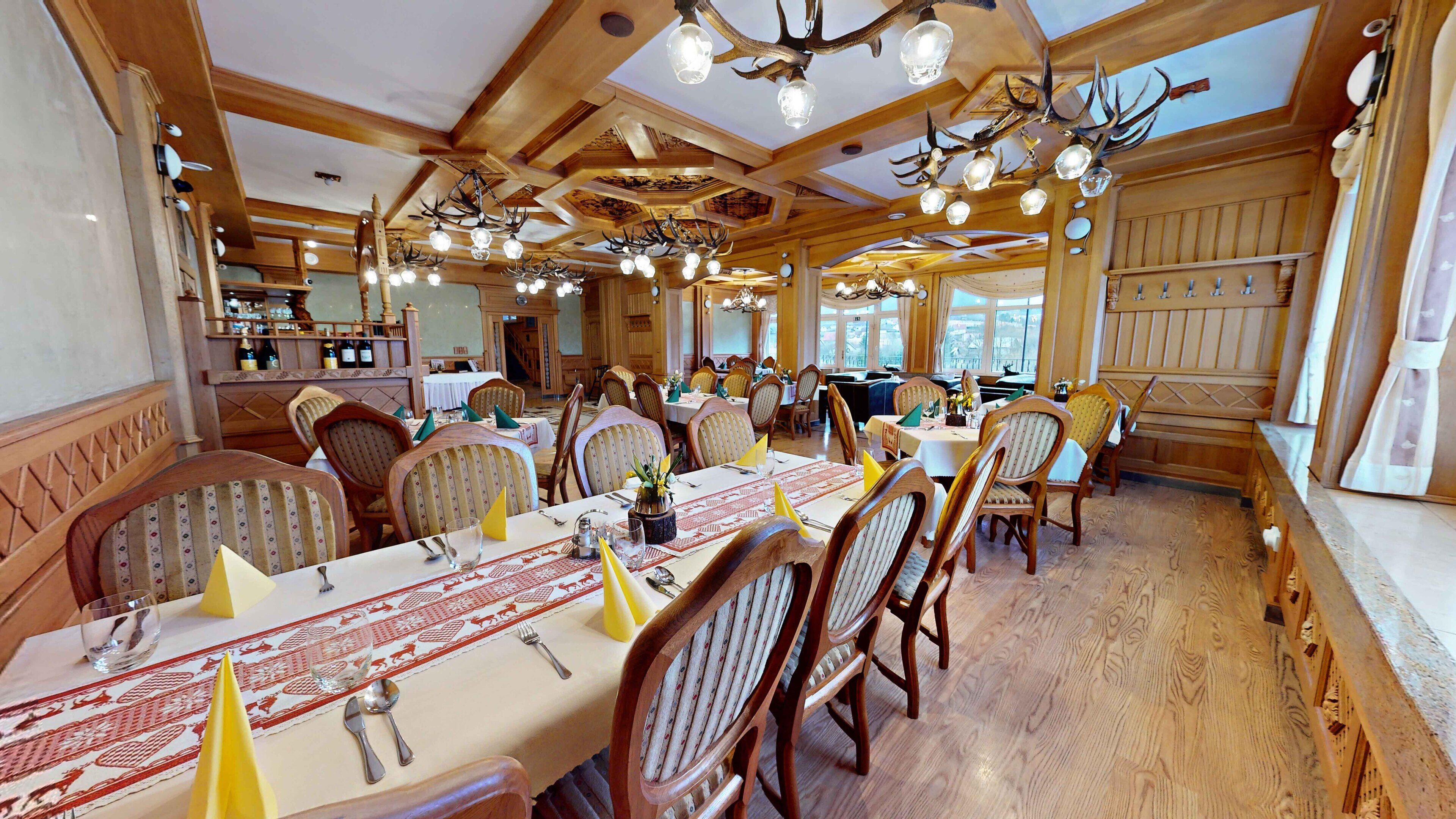 A dining area with wooden tables and chairs, chandeliers with antler decorations, and a bar area in the background.