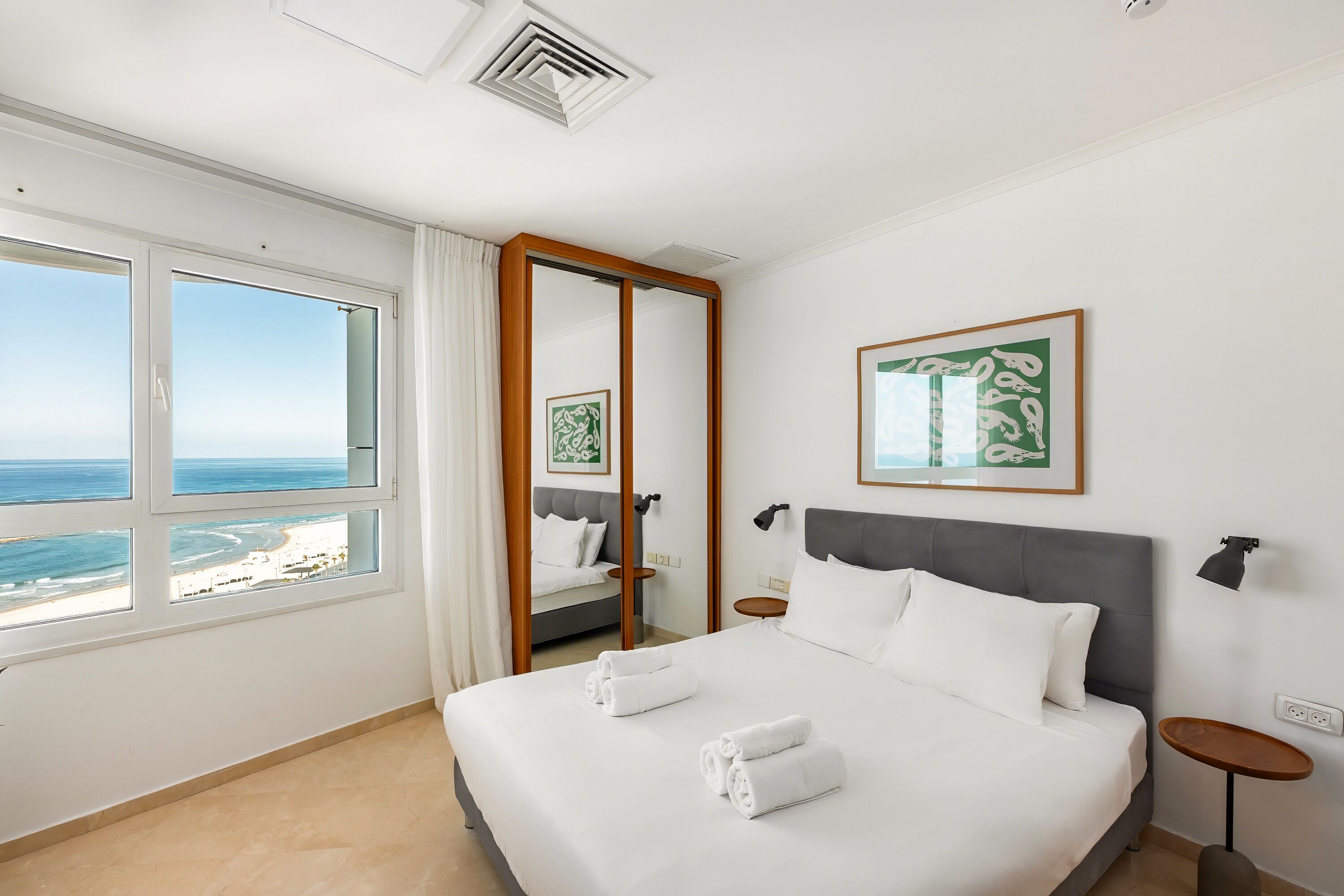A hotel room with a large bed, a view of the sea, a mirror, and a framed picture on the wall.