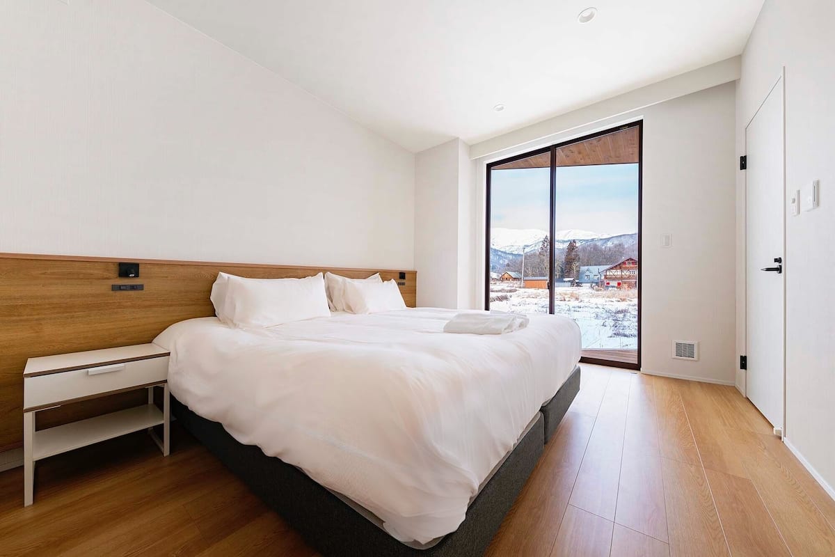 A modern hotel room with a large bed, a bedside table, and a view of a snowy landscape through a sliding door.