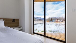 Un dormitorio con un amplio ventanal corredero que da a un espacio exterior nevado y vistas a montañas y edificios.