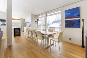A modern kitchen with white cabinets, a dining area with a wooden table and chairs, and large windows offering a view of the outdoors.