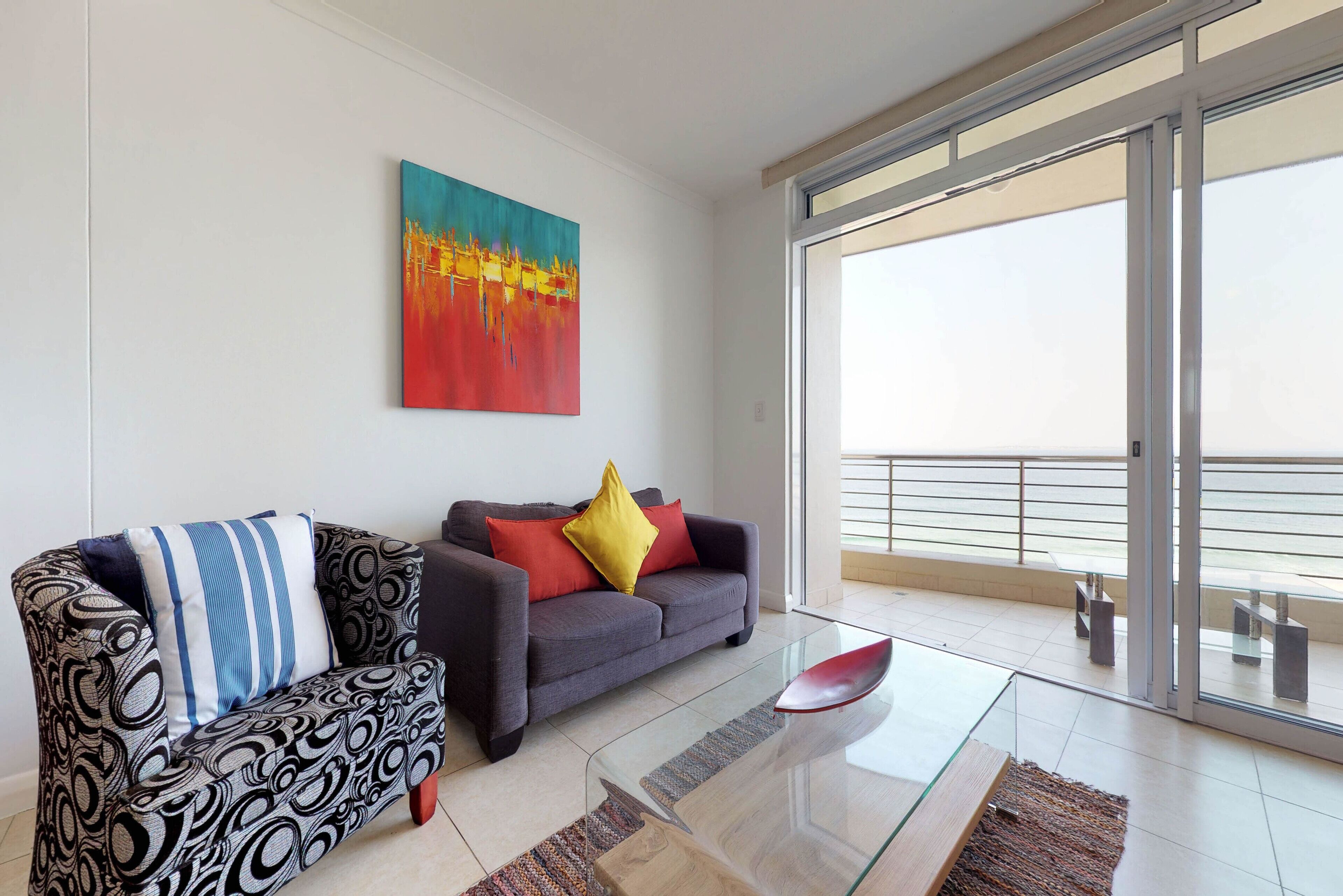 A modern living room with a grey sofa, a patterned armchair, and a glass coffee table. A colorful abstract painting is on the wall, and the room has a large sliding glass door leading to a balcony.