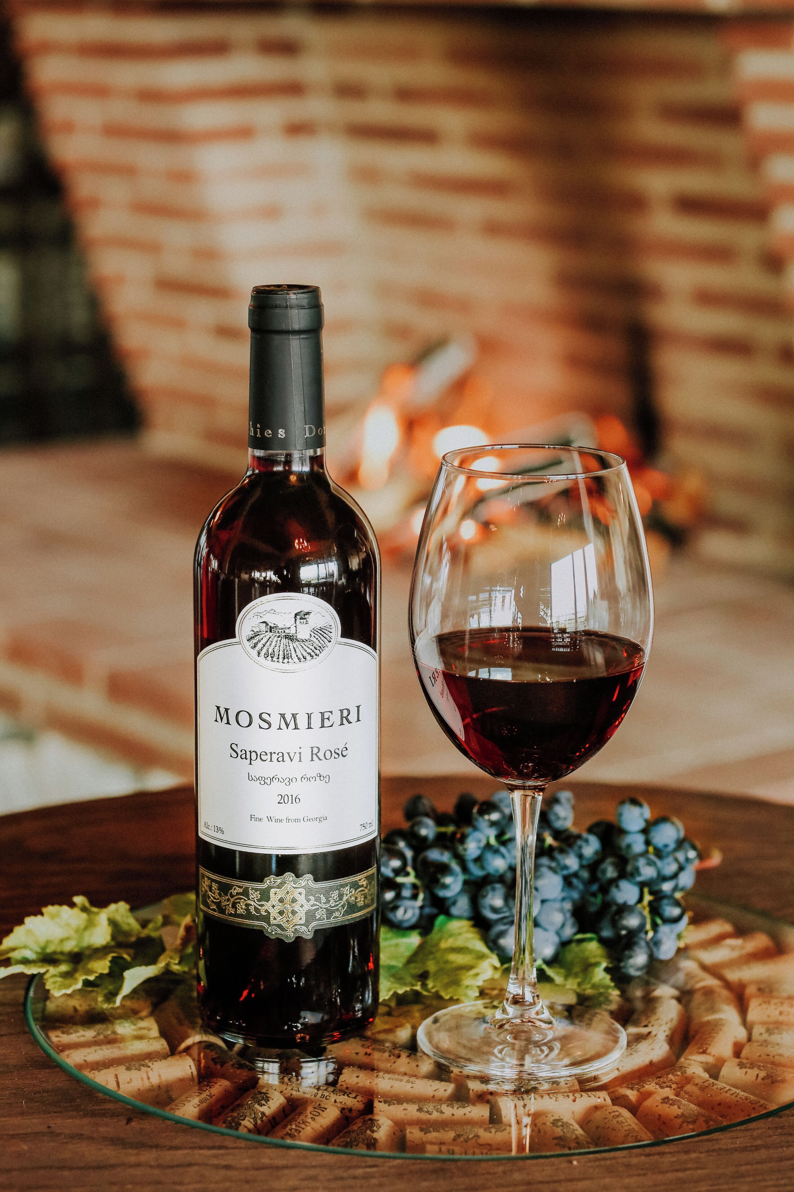 A bottle of Mosmieri Saperavi Rosé wine and a glass of red wine on a table with a glass plate, surrounded by grapes and corks.