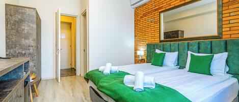 A hotel room with a brick wall, a large bed with green bedding, a wooden dresser, and a green patterned rug.