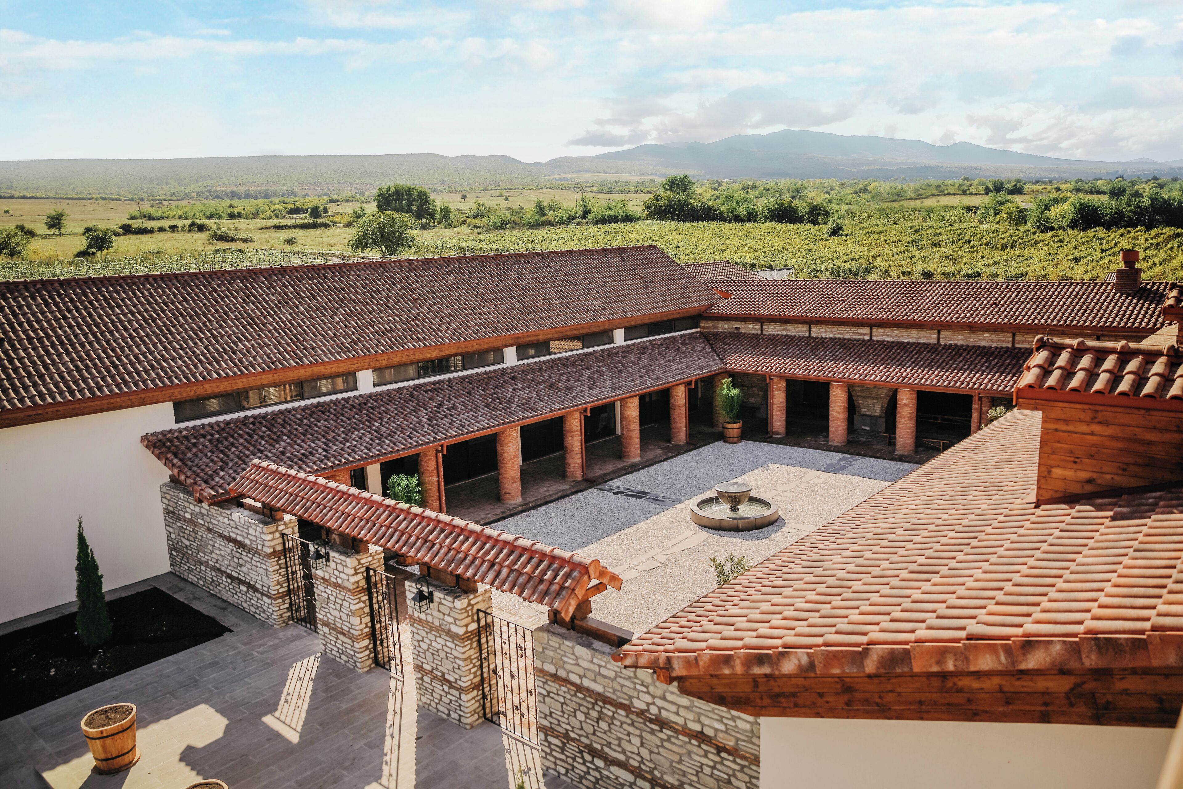 A courtyard with a tiled roof, stone walls, and a view of a vineyard and mountains.