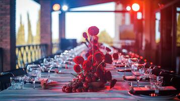 A long dining table set with red flowers, glasses, and plates, in a dimly lit room with a brick wall and a large screen in the background.