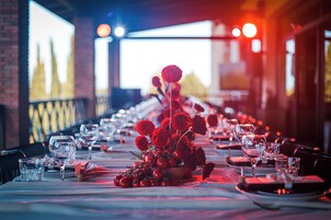 A long dining table set with red flowers, glasses, and plates, in a dimly lit room with a brick wall and a large screen in the background.