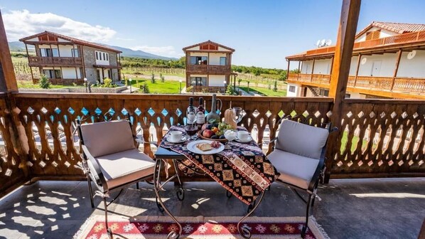 A balcony with a table set for two, overlooking a vineyard and two buildings. - Château Mosmieri Hotel & Winery (Telavi)
