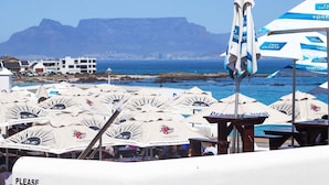 Una zona sulla spiaggia con diversi ombrelloni e tavoli, vista sull'oceano e sulle montagne sullo sfondo, e un cartello con la scritta 'PLEASE'.