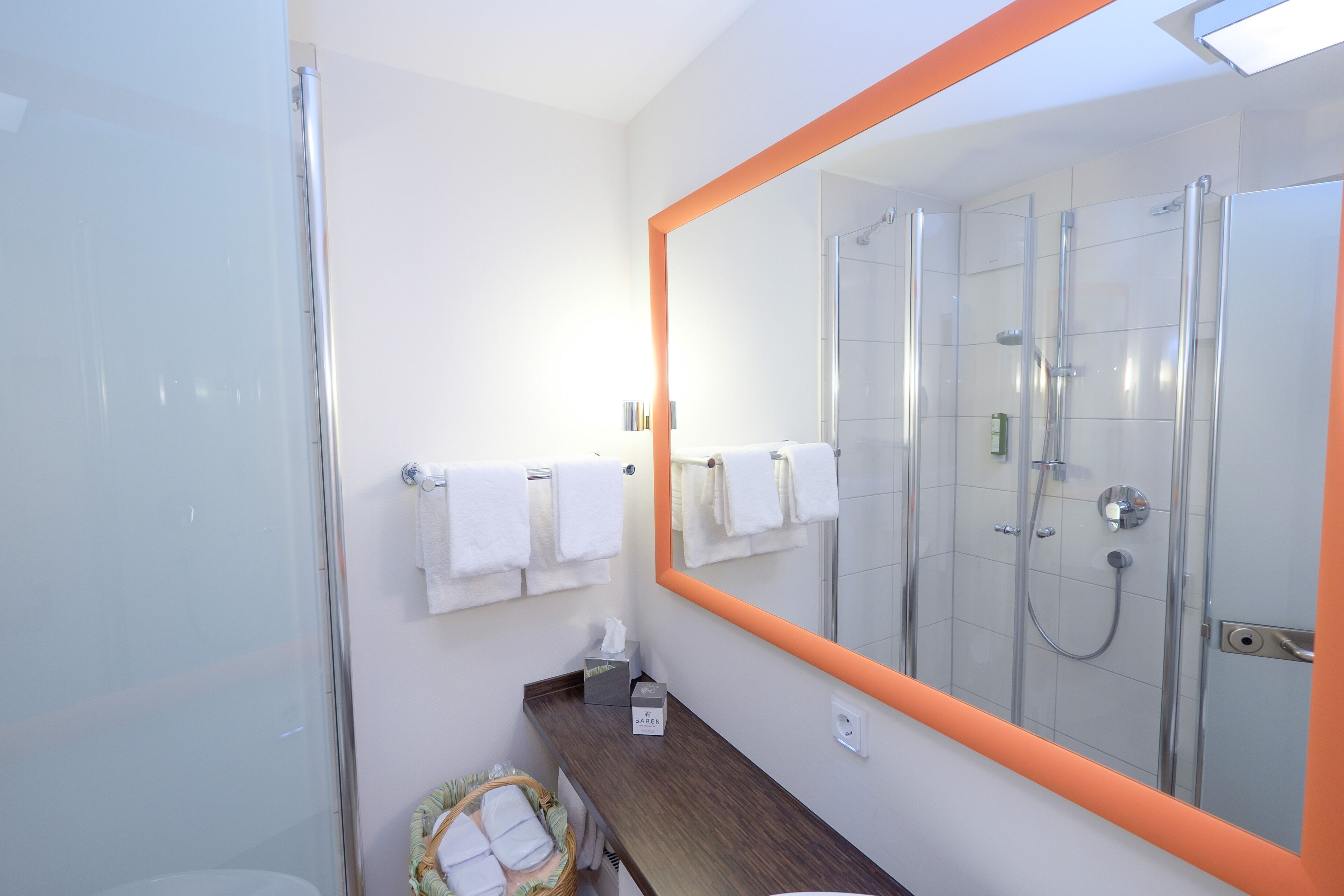 A modern bathroom with a glass shower enclosure, a large mirror with an orange frame, and a wooden vanity.