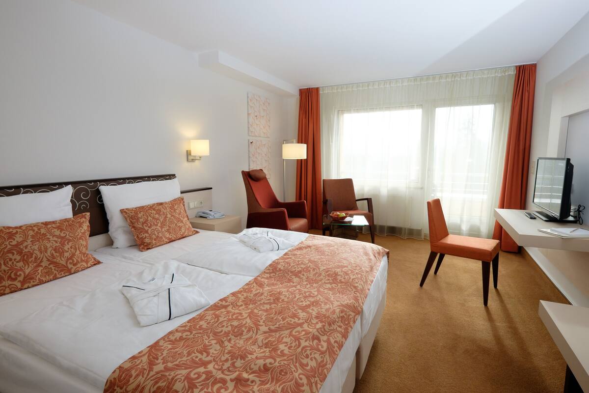 A hotel room with a bed, two chairs, a desk, a television, and a window with curtains.