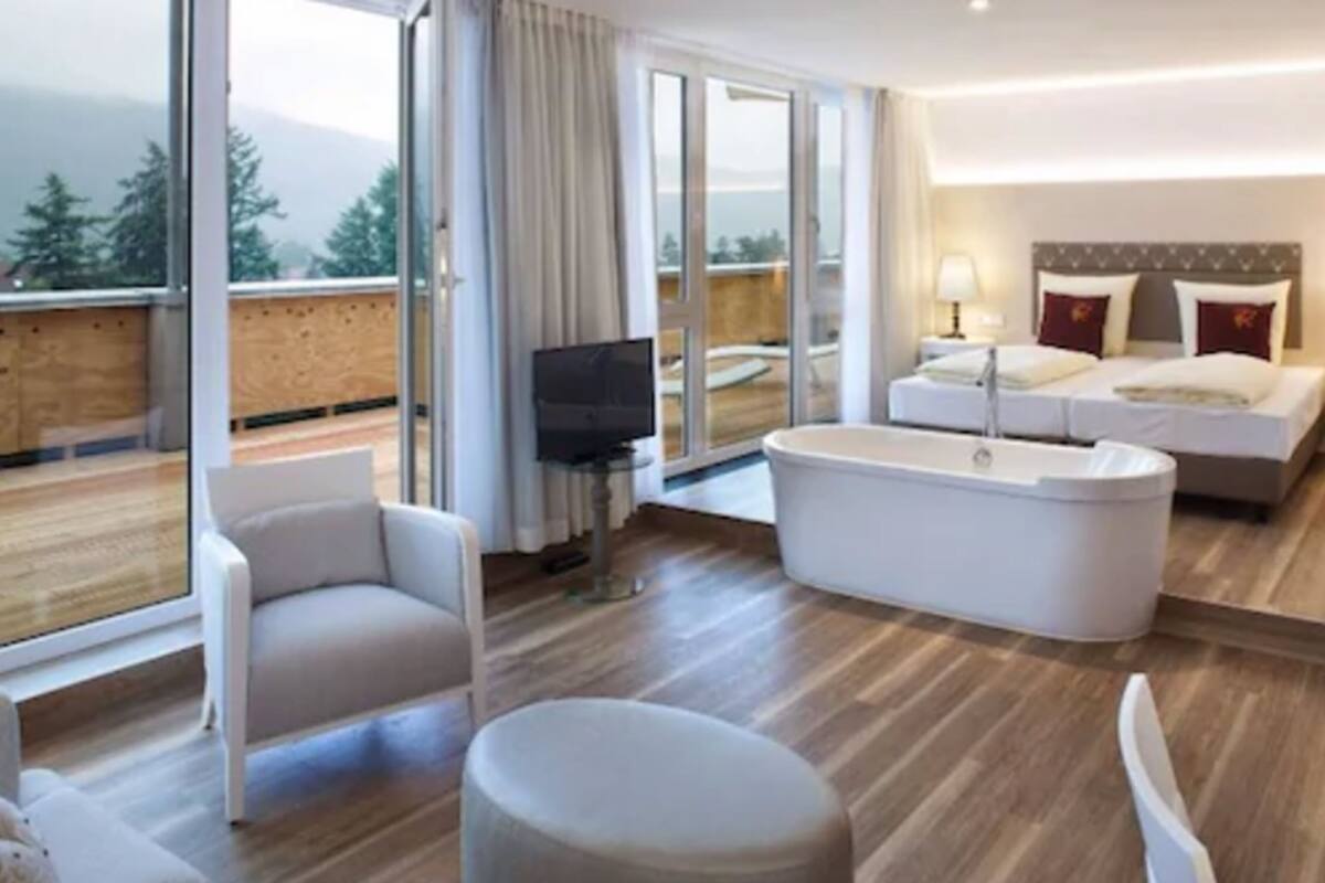 A modern hotel room with a large bed, a sofa, a TV, and a view of the outdoors.
