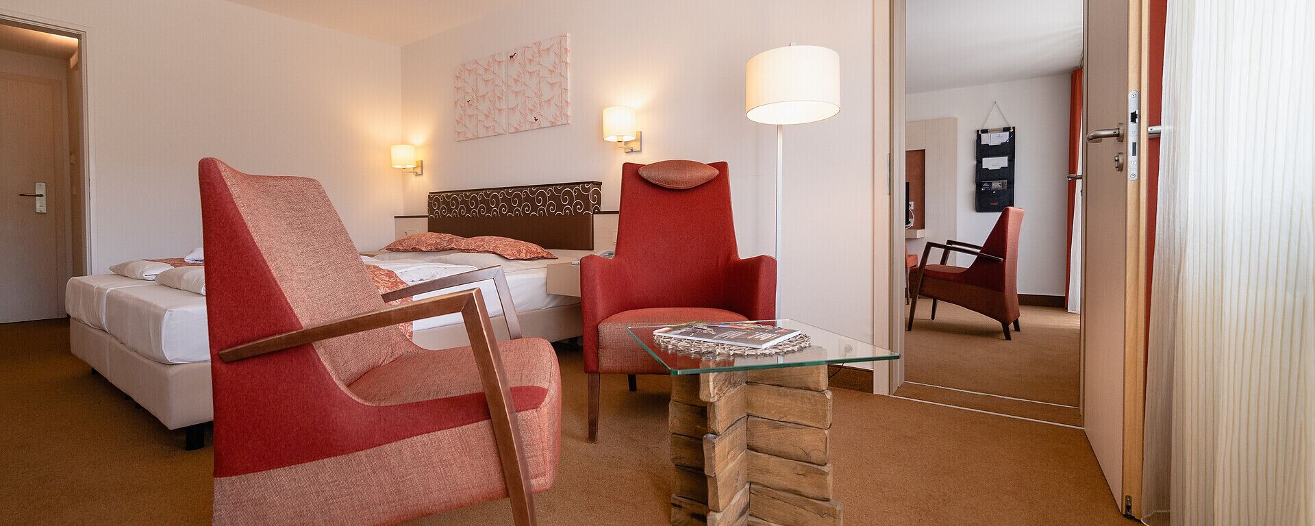 Ein Hotelzimmer mit einem Bett, zwei roten Sesseln, einem Glastisch und einem Wandleuchten sowie einem Gemälde.