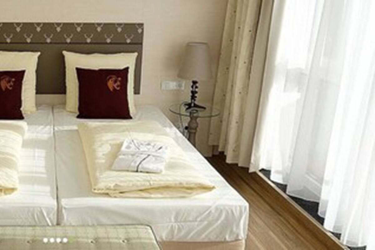 A double bed with white linens and two pillows, a bedside table with a lamp, and a window with curtains.