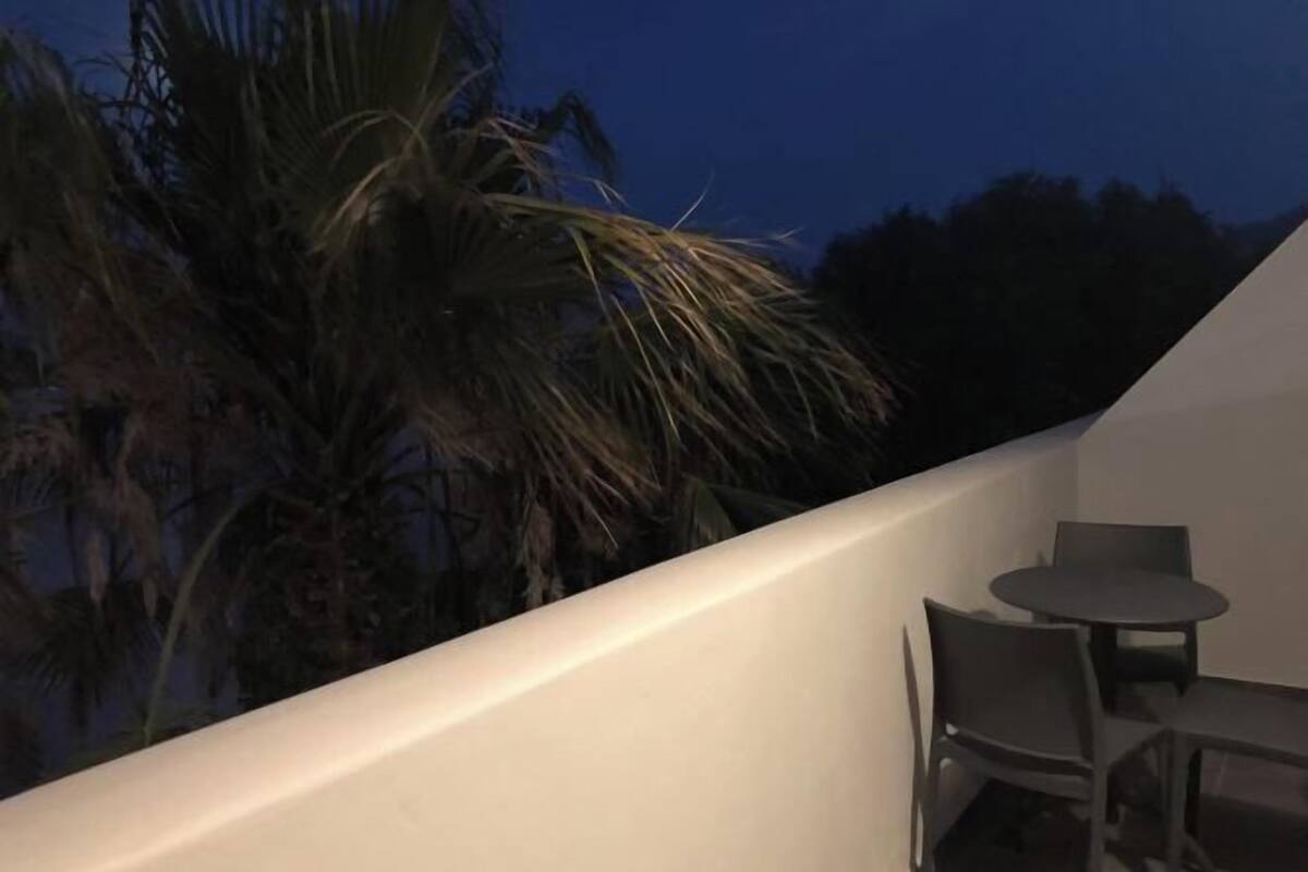 A balcony with a table and chairs, overlooking palm trees and a dark sky.