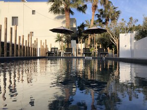 A pool with chairs and umbrellas, surrounded by a wooden fence and palm trees. - Dragonfly Villas (Santorini)