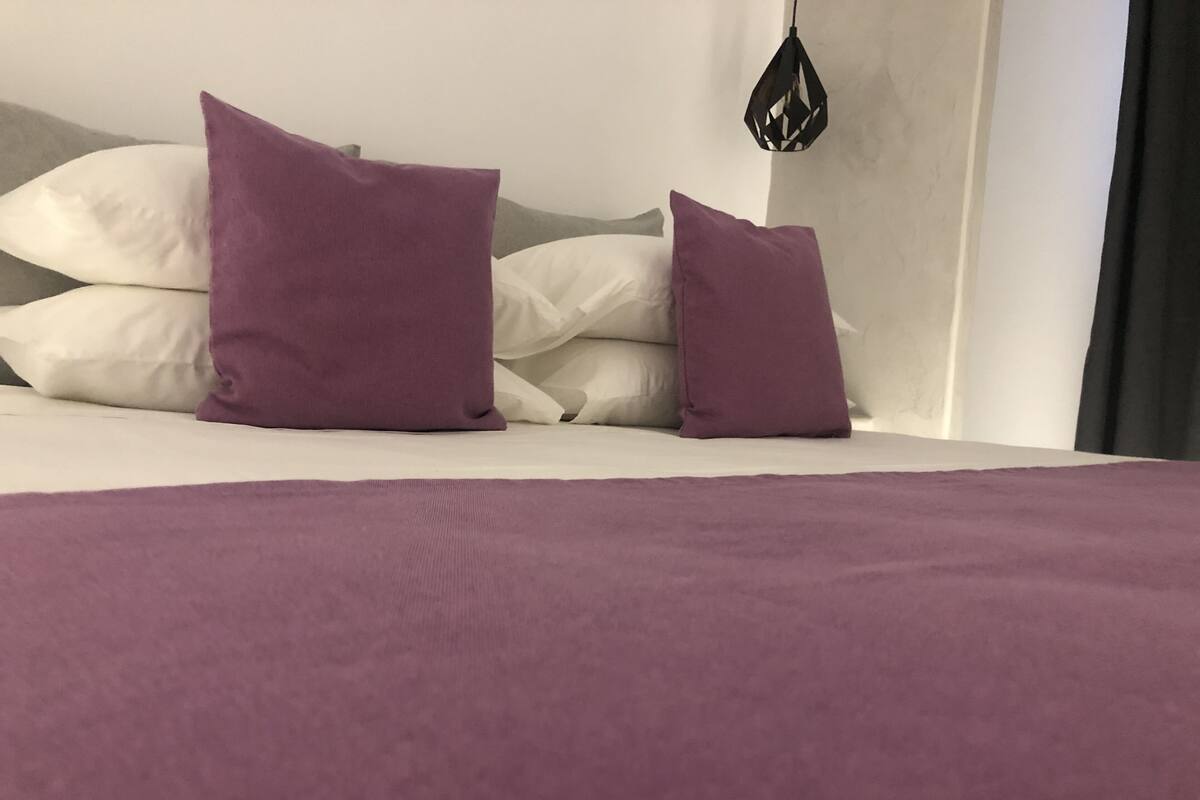 A bed with a white mattress and a purple blanket. There are two purple pillows and a white pillow on the bed.